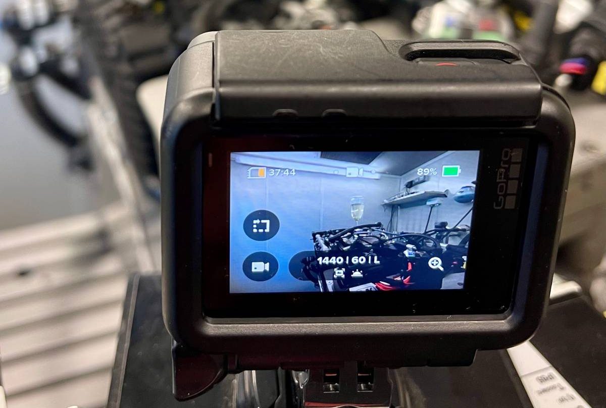 Im Vordergrund des Bildes ist ein Monitor zu sehen: Auf dem sogenannten „Champagner-Motor“ von Obrist steht ein Glas mit Champagner. Der kompakte Zweizylindermotor läuft vibrationsfreier als ein Zwölfzylinder, was das Glas Champagner beweist.