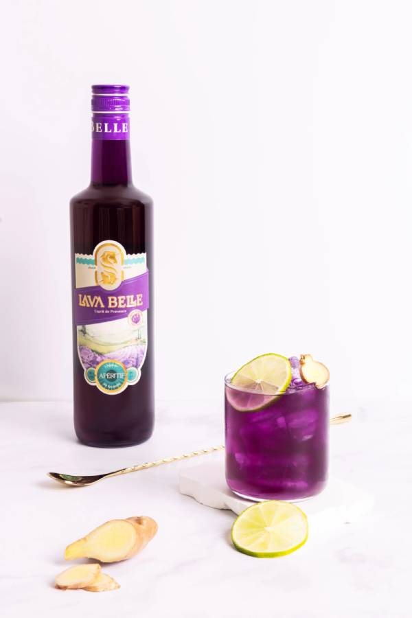 Lav'a Belle Nizza Mule alkoholfrei - Lila Flasche, ein lila Glas mit Eis und Zitrone, ein goldener Löffel und andere Accessoires.