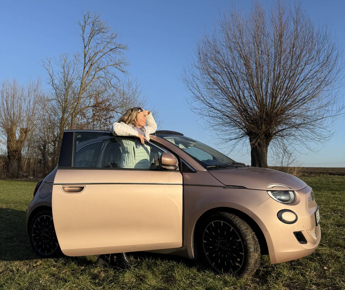 Eine junge schöne Frau steht hinter der geöffneten rechten Tür ihres FIAT 500e Cabrio und schaut nach vorne.