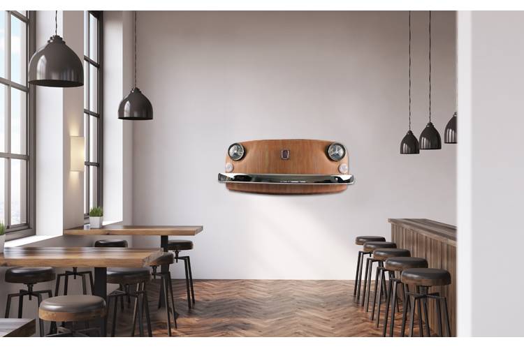 Eine Frontmaske hängt an einer Wand in einem Raum mit 2 rustikalen Tischen, fünf Hockern an einer Bar und einigen Hängelampen in Metalloptik.