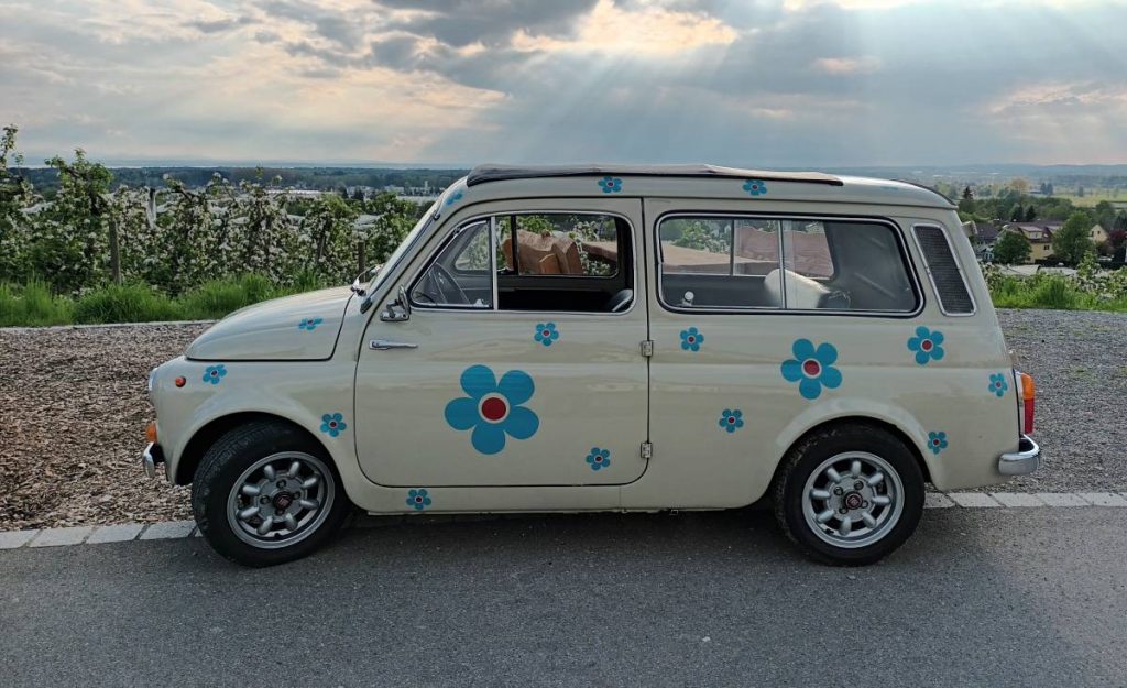 Ein Oldtimer Fiat Giardinierea in der Farbe Weiß mit blauen Blüten auf dem Lack und mit geschlossenem Schiebedach steht vor einer lichtdurchfluteten Landschaft mit blühenden Obstbäumen.