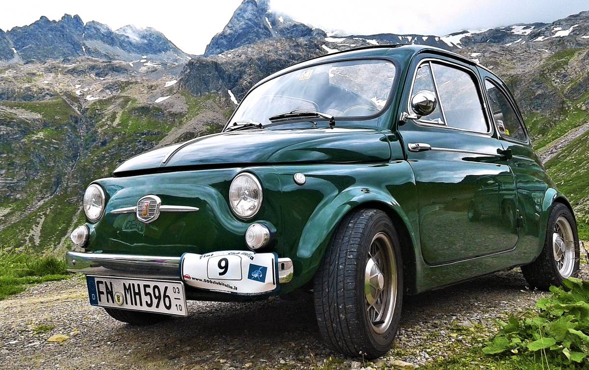 Ein Oldtimer Fiat 500 in der klassischen Farbe Dunkelgrün parkt hoch oben inmitten von schroffen Bergen. Das Fahrzeug ist von schräg unten links in Großaufnahme zu sehen.