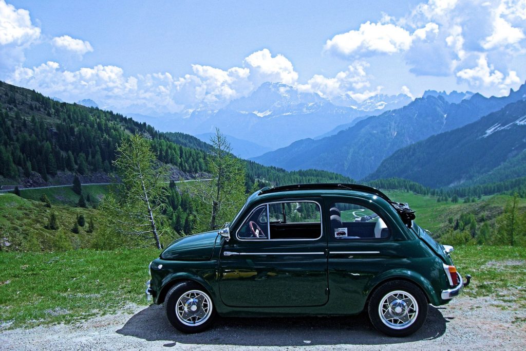 Ein Oldtimer Fiat 500 in der klassischen Farbe Farbe Dunkelgrün und mit geöffnetem Schiebedach steht vor einer imposanten Bergkulisse.