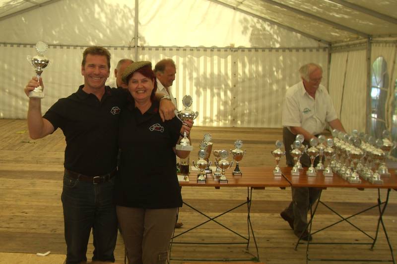 Ein Mann und eine Frau zeigen stolz ihre Pokale. Daneben sind noch viele weitere silberne Pokale auf einem Tisch zu sehen.